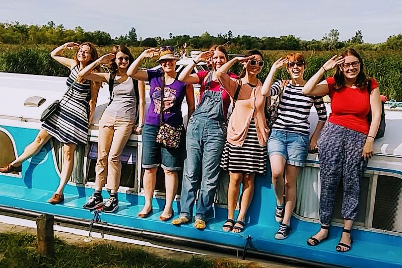 A group of seven women (five are uklibchat team members) standing on the side of a boat on the Norfolk broads. All are smiling and saluting the camera.